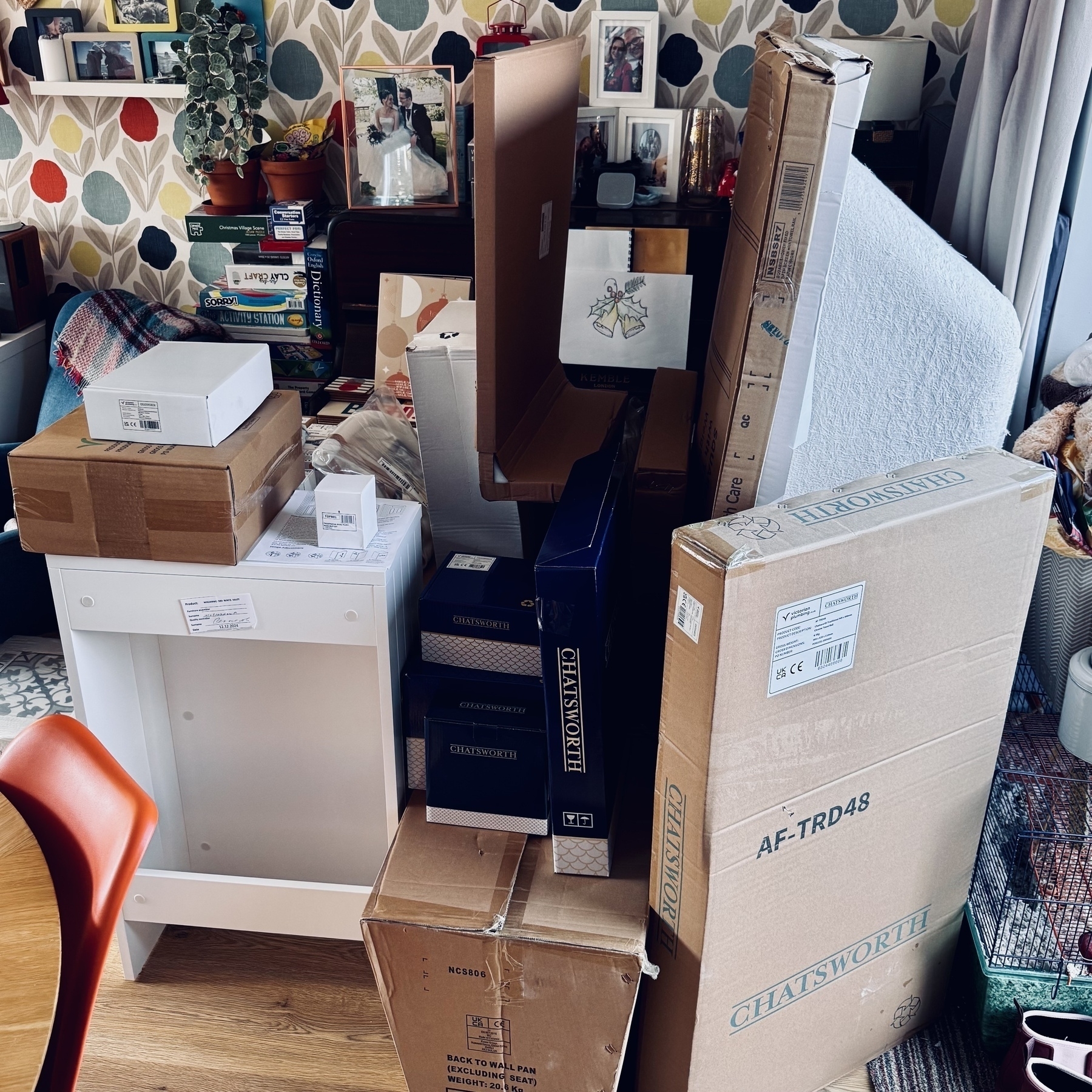 A room filled with stacked cardboard boxes, various packages, and furniture against a colourful, patterned wallpaper.