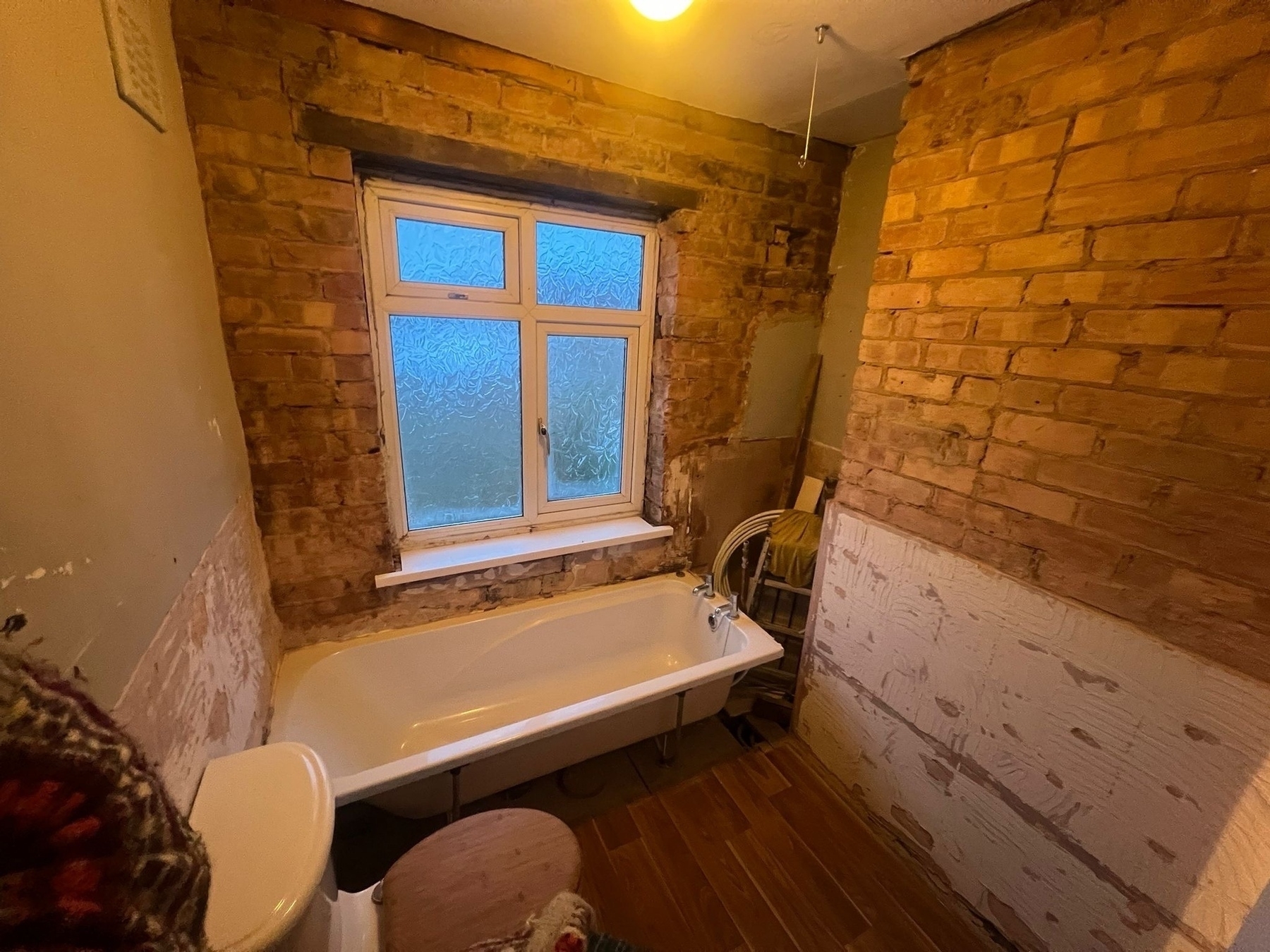 Auto-generated description: A bathroom under renovation features exposed brick walls, a bathtub, a toilet, and a frosted window.